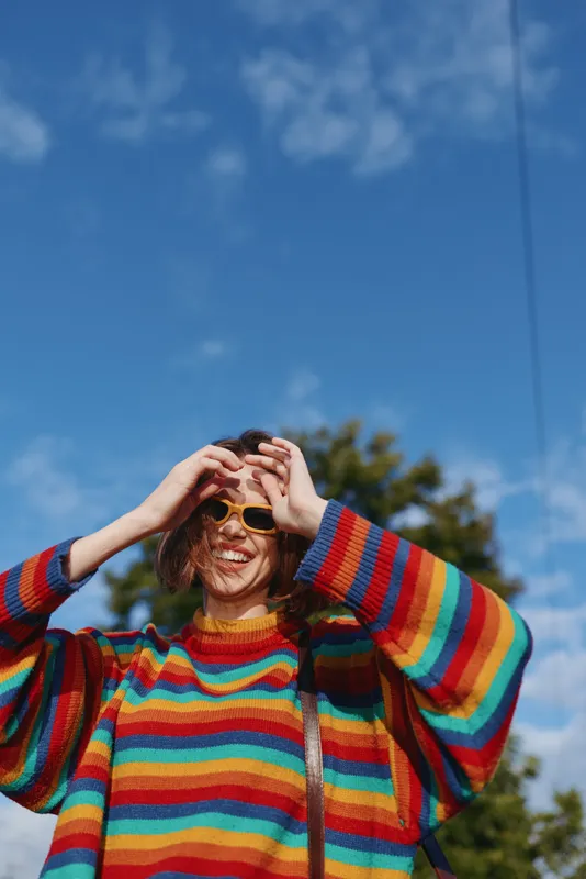 Young woman in a colorful striped sweater and yellow sunglasses, smiling and adjusting glasses outdoors under a clear blue sky. Curly hair, playful lifestyle portrait for fashion and travel imagery.
