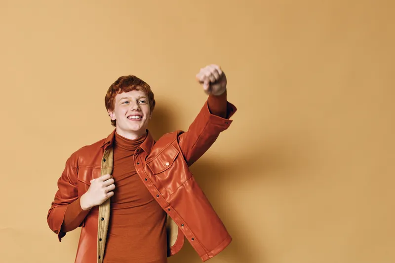 Young man with red hair wearing orange leather jacket and turtleneck posing with fist raised, smiling confidently against plain brown background in studio portrait.