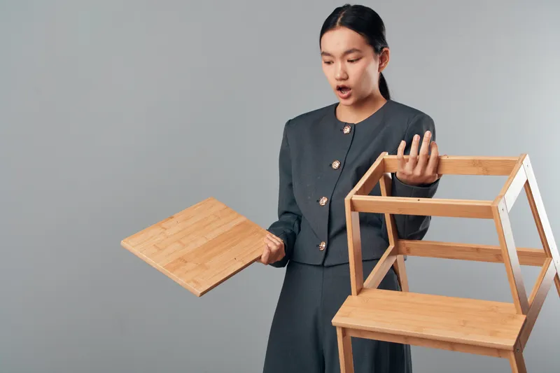 Woman with surprised expression holding wooden shelf parts indoors, assembling furniture with focus on wood pieces and hands in casual clothing against plain background.