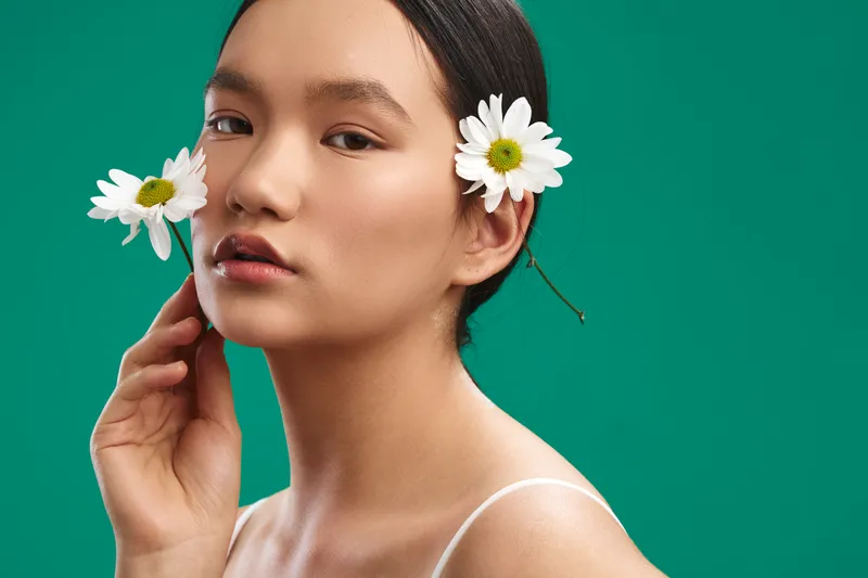 asian woman with daisies in her hair stands against a vivid green backdrop, a soft portrait that celebrates natural beauty, calm confidence, and a light, fresh contemporary vibe.