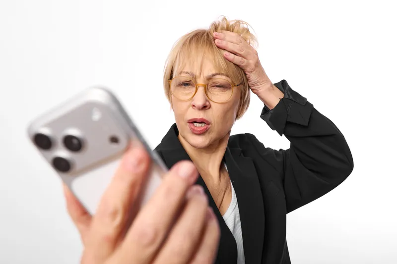 Woman stress smartphone glasses older selfie headache panic, she covers her head and looks worried, holding mobile for a selfie, indoors office, white background
