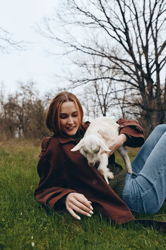 Young woman with braided hair lies on grass holding a white baby goat in her arms during a relaxed outdoor lifestyle moment in early spring.