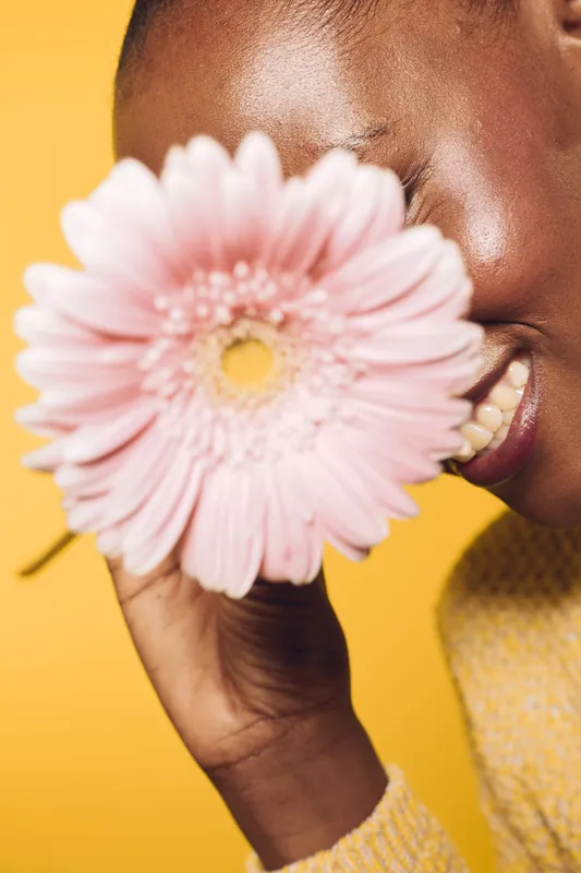 Close-up of happy dark skinned African woman smiling and holding pink flower near face on yellow background. Concept of natural beauty, joy, skincare, and floral wellness.