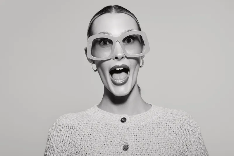 Surprised white woman with fair skin and slicked back hair wears oversized glasses and knitted sweater, posing on light background. Concept for fashion, emotion, and eyewear.