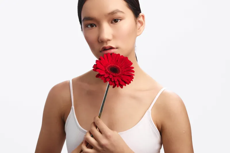 asian woman holds a red flower close to her lips in a simple studio portrait. soft lighting highlights smooth skin and calm expression, creating a warm, intimate mood.