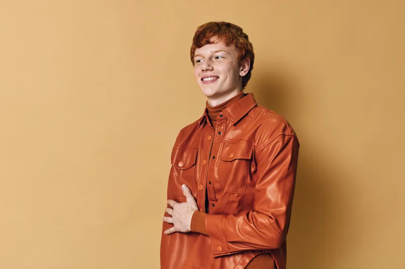 Young man with red hair smiling and wearing brown leather jacket posing against beige background in studio. Portrait of casual male model with modern hairstyle and confident look.