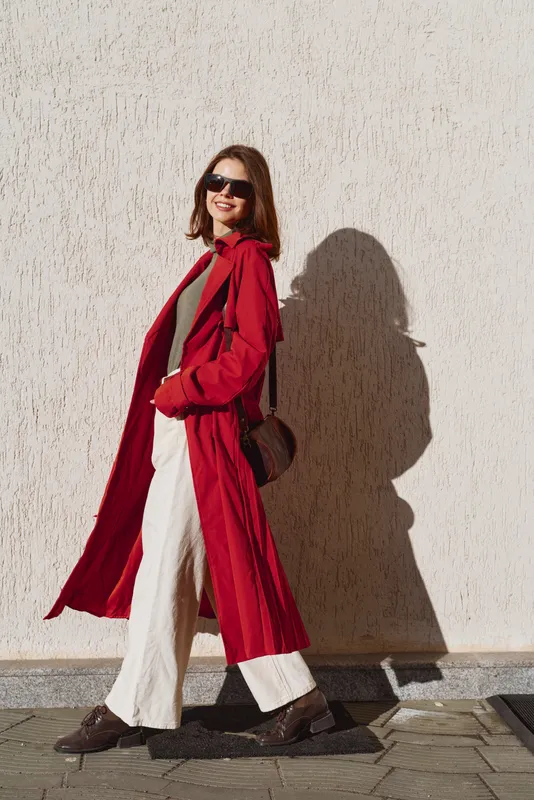 Young woman in a long red coat walks confidently along a sunlit wall wearing sunglasses and white pants, enjoying a stylish autumn day outdoors.