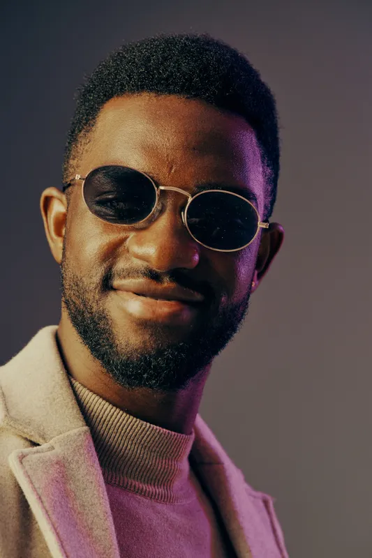 Stylish male model wearing sunglasses and a coat, posing confidently against a colorful backdrop, expressing sophistication and modern fashion trends