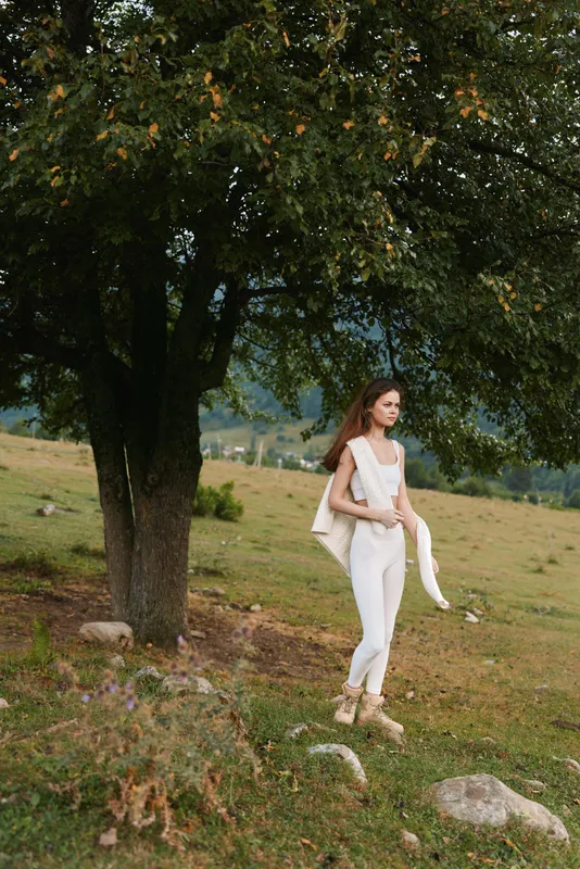 Young woman in white outfit walks near large tree in open field holding a white jacket over her shoulder enjoying nature and peaceful outdoor lifestyle.