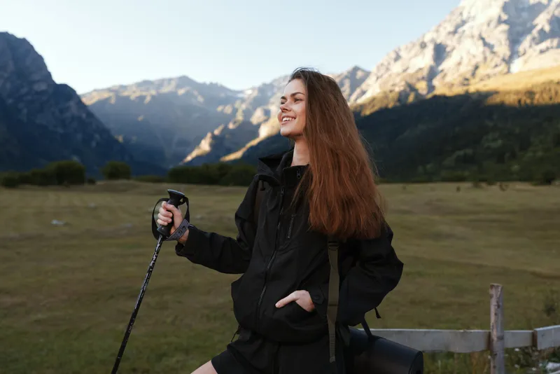 A cheerful woman outdoors stands in a mountain valley, smiling as she grips a trekking pole. The scene features rugged peaks, green meadows, and clear skies, creating an uplifting mood of adventure