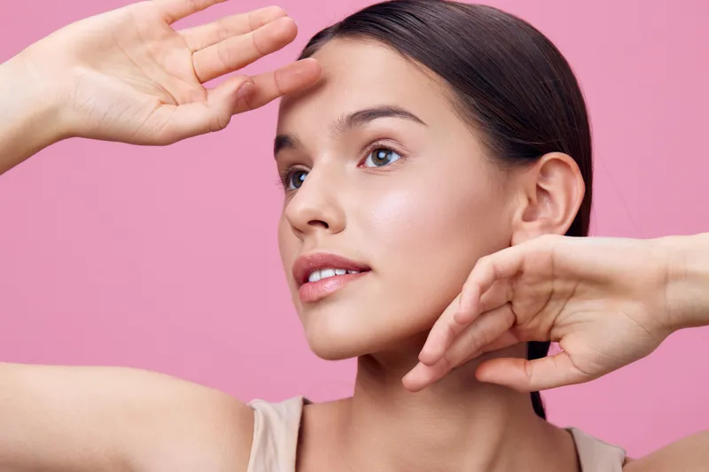 Young woman with natural makeup poses against a pink background, showcasing a serene expression and elegant hand movements, perfect for beauty and skincare themes