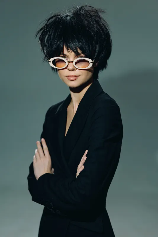 Stylish woman with short black hair and white sunglasses wearing a black blazer, posing confidently against a neutral background.