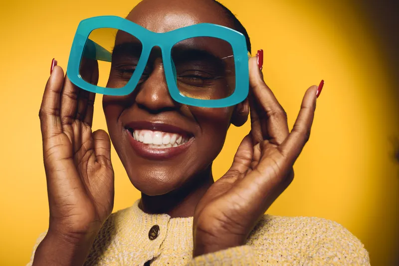 Happy African woman with dark skin wearing large blue glasses and yellow sweater smiles with closed eyes on yellow background. Concept of joy, fashion, and beauty portrait.