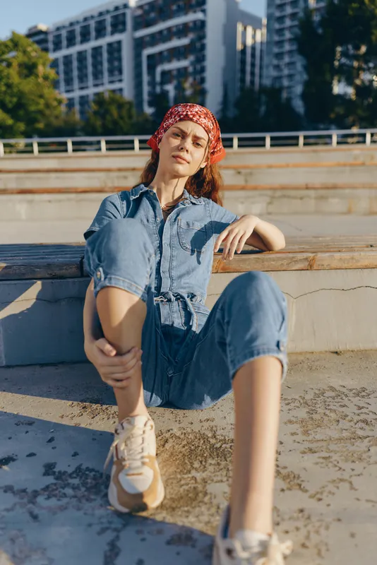 Lifestyle woman in modern boho-western denim street style sitting relaxed outdoors, wearing casual sneakers and red bandana under warm natural light film color.