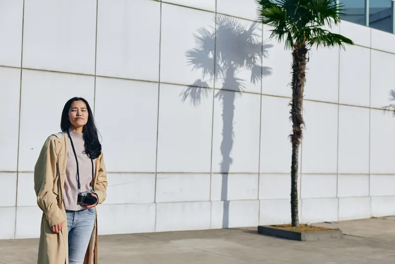 Woman with camera outdoors near a white wall, wearing a beige coat and jeans, palm tree shadow creates a striking backdrop for casual street photography.