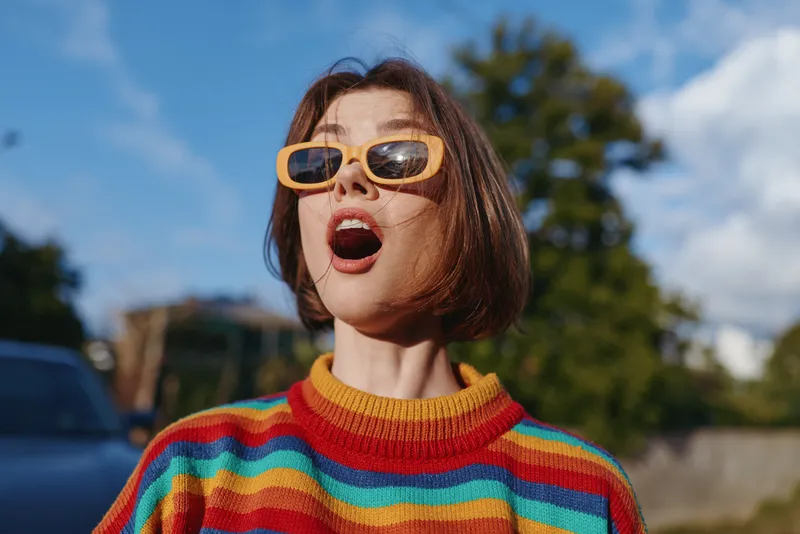 Woman in striped sweater and yellow sunglasses with surprised expression, short bob hairstyle outdoors on a sunny day street portrait, colorful retro knit lifestyle and fashion vibe.