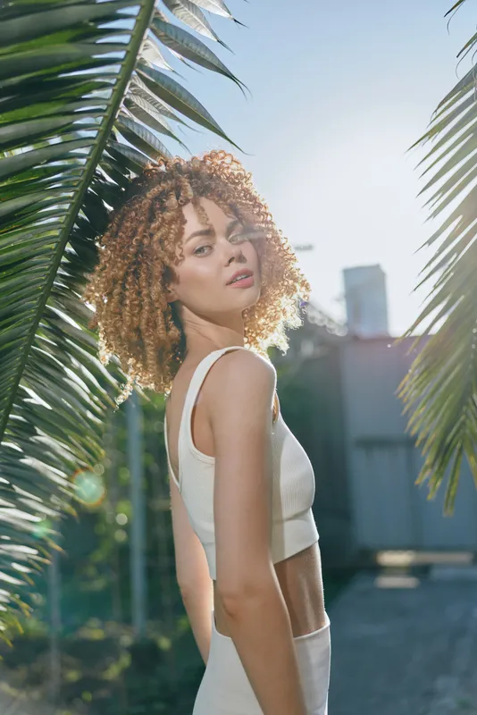 Young woman with curly hair wearing white top stands outdoors between green palm leaves under bright sunlight, showcasing natural beauty and a calm, confident expression.