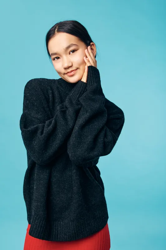 Young woman with black hair wearing oversized dark sweater and red skirt posing with hand on face against blue background. Portrait of smiling female in casual warm clothing with relaxed expression.