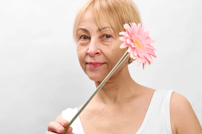 Elderly woman portrait holding a pink flower on a white background closeup soft light gentle smile relaxed pose minimal scene copy space calm mood side view