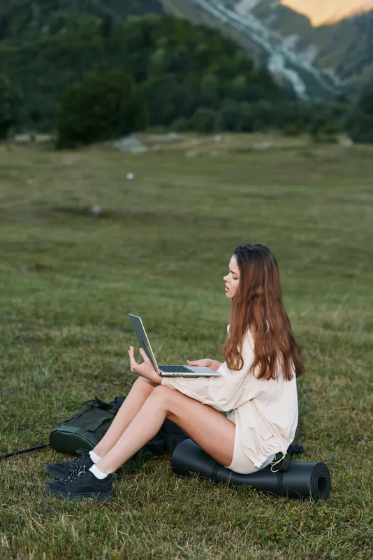 A woman sits on a grassy field typing on a laptop with a focused expression, enjoying a calm landscape and fresh air in a natural outdoor workspace.