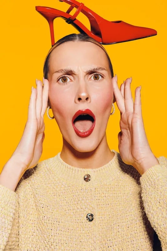 Surprised woman with red lipstick and earrings balances red high heel shoe on her head against bright yellow background. Concept for fashion, style, and playful expression.