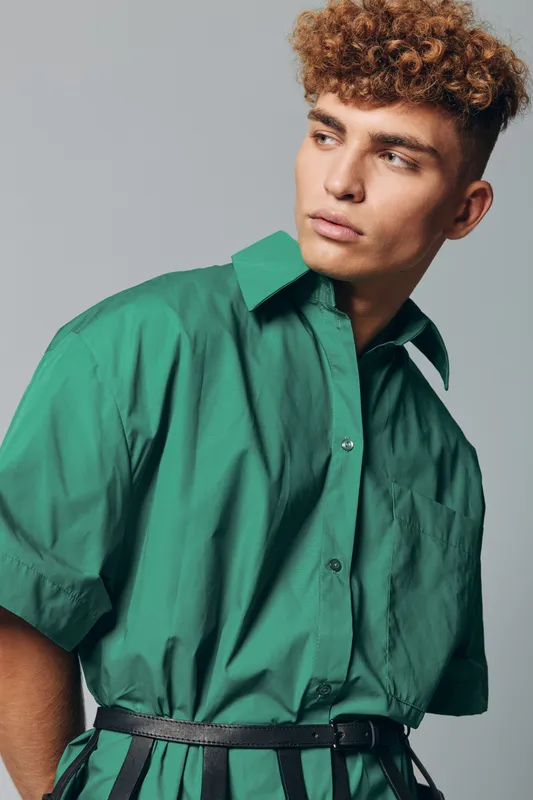 Young man with curly hair wearing green shirt and black belt posing against gray background looking sideways with serious expression in studio portrait.