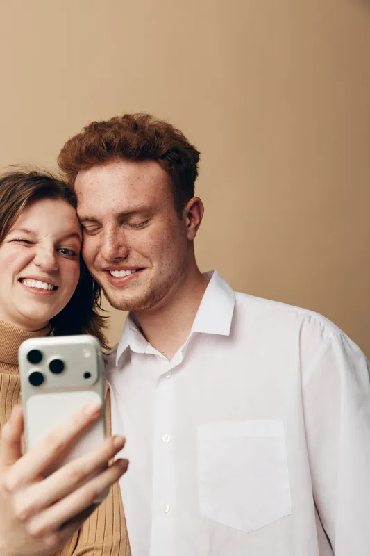 Young couple taking a selfie with smartphone, smiling and enjoying moment together. Casual clothing and warm beige background create cozy and intimate atmosphere of friendship and love.