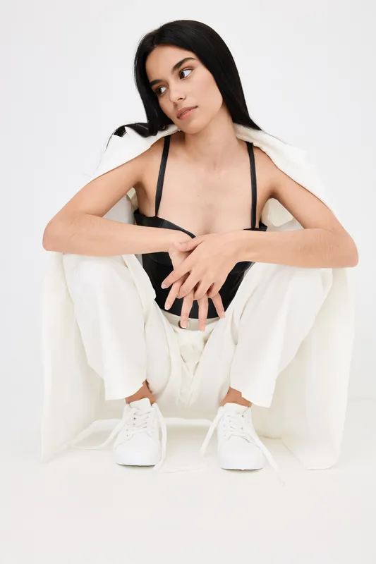 young woman in black top and white pants sitting on floor with thoughtful expression, long dark hair, minimalist studio background, casual fashion style