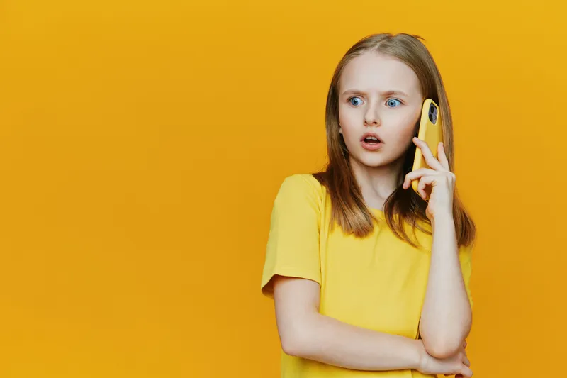 Surprised girl in a yellow shirt talking on the phone against a vibrant yellow background, expressing shock and curiosity, perfect for communication themes