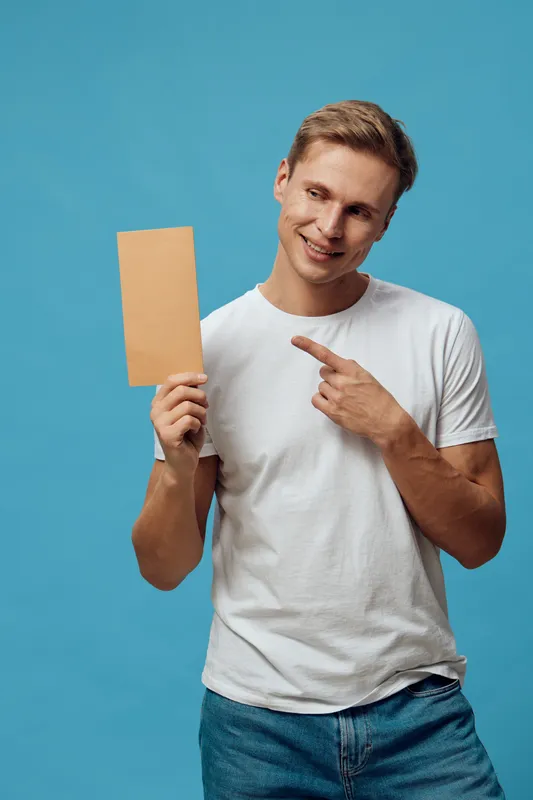 Smiling man holding blank cardboard sign and pointing at it isolated on a bright blue background, casual clothing, cheerful mood, studio portrait, lifestyle concept.