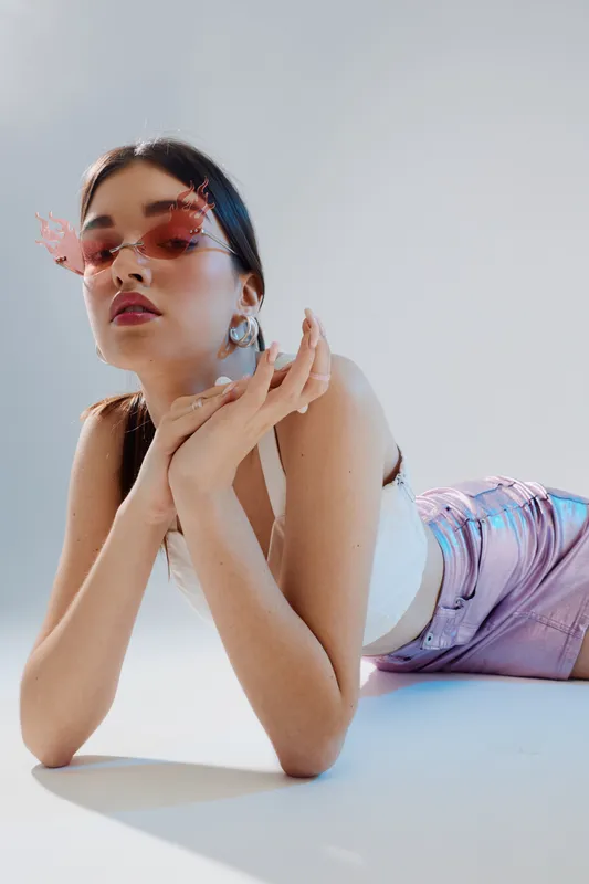 Young woman in trendy pink sunglasses and shiny shorts poses lying down on white floor. Studio shot with soft lighting. Concept of woman beauty fashion and modern style.