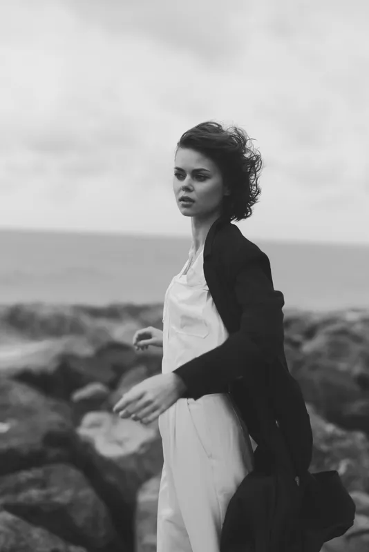 Woman in a dress and coat on a windy shoreline among rocks, sea in the background, monochrome mood with dramatic atmosphere and coastal movement