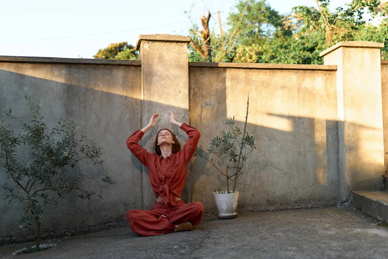 Woman outdoors in a red outfit sits cross legged on a paved surface, hands raised above her head in a yoga inspired pose, calm smile, sunlit scene, natural colors lifestyle outdoor yoga meditation
