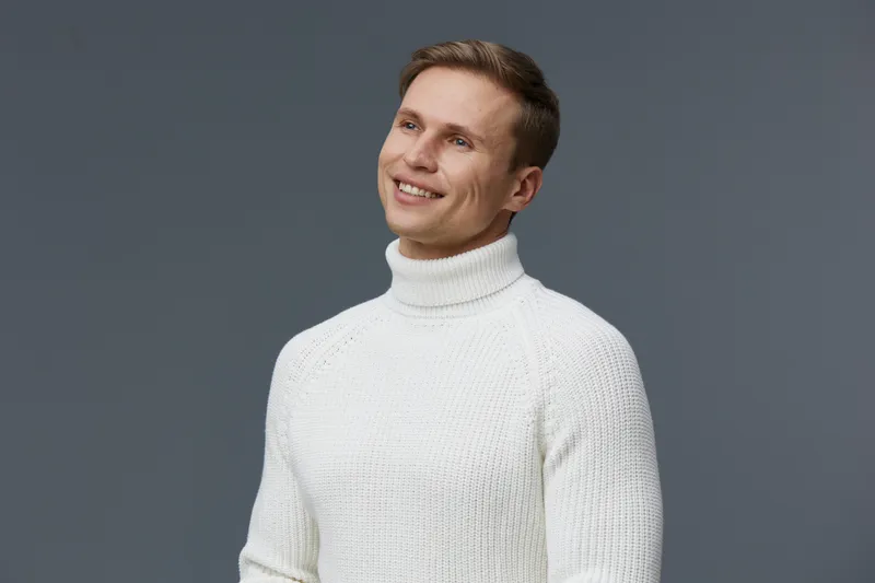 Smiling man wearing white turtleneck sweater on gray background, happy expression, casual style, studio shot, cheerful mood, fashion look, portrait.