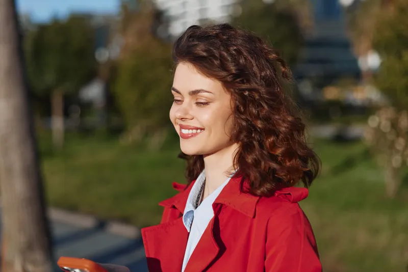 Young woman with curly hair wearing red coat smiles while looking at smartphone outdoors in sunny park, enjoying positive mood and casual style.