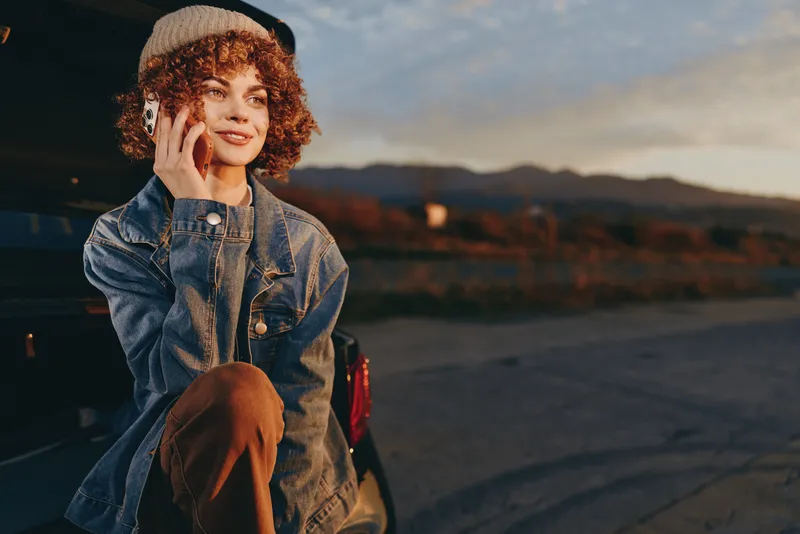 Woman lifestyle portrait outdoors at sunset, curly haired woman in denim jacket holds phone to ear, smiling on a quiet road parking area, calm communication evening travel vibe.