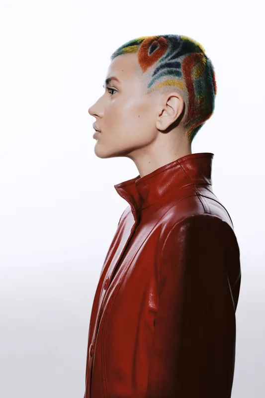 Woman with colorful patterned short hair wearing a red leather jacket stands in profile against a white background, showcasing bold, artistic hairstyle and modern fashion.