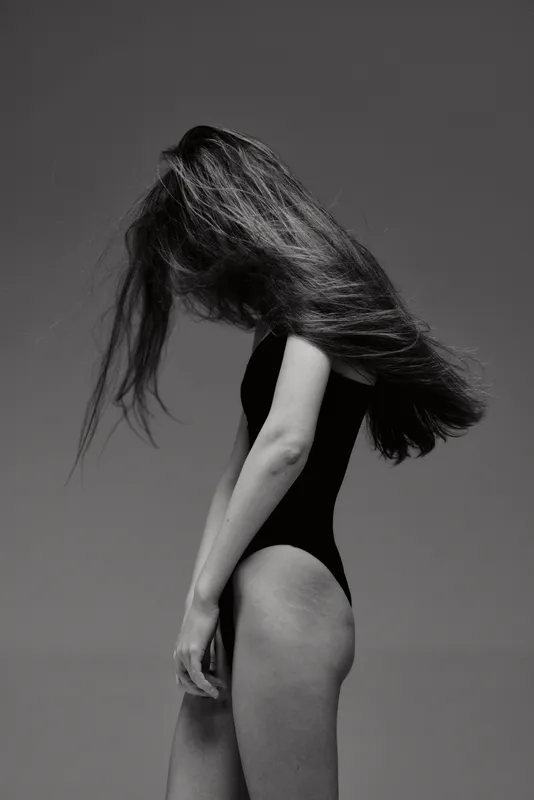 Black and white portrait of a woman with long hair wearing a black bodysuit against dark background.