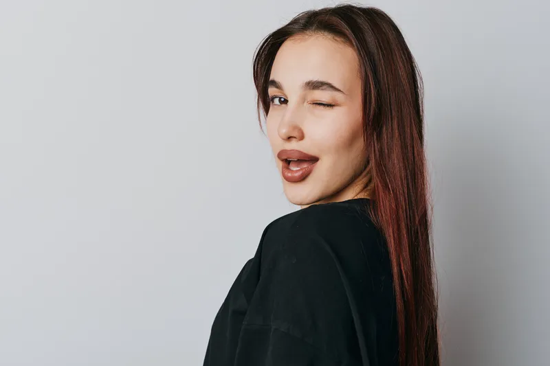 Young woman with long hair wearing black shirt winking with lips slightly open on gray background. Portrait of attractive female with confident expression in studio lighting.