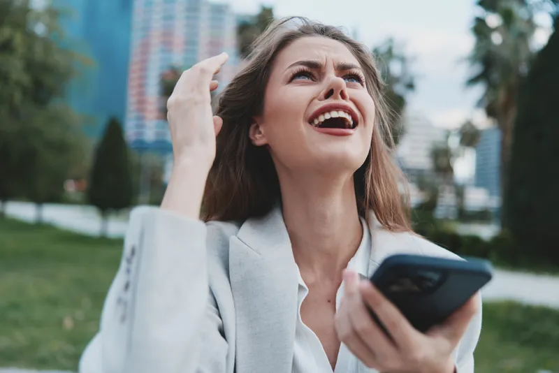 Emotional woman in business attire frustrated while using smartphone in outdoor urban setting with greenery and modern buildings in background.