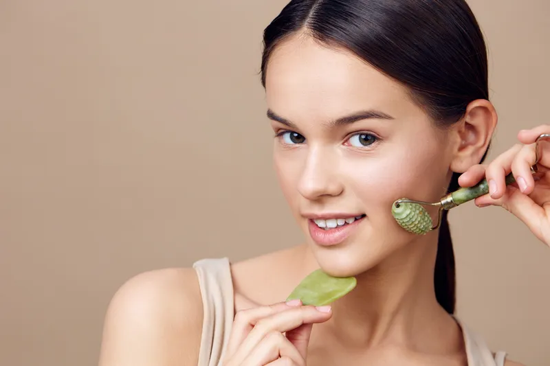 Young woman using a jade roller for facial massage in a beauty routine, showcasing healthy skin and a natural glow in a soft, neutral background