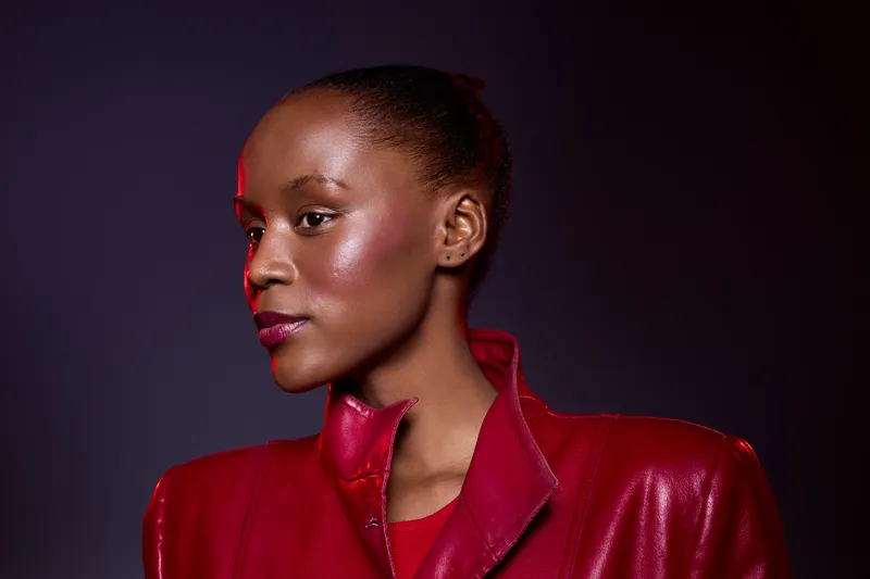 Portrait of a confident black woman with dark skin wearing a red leather jacket against a dark purple background. Concept of fashion, elegance, and beauty with strong lighting.