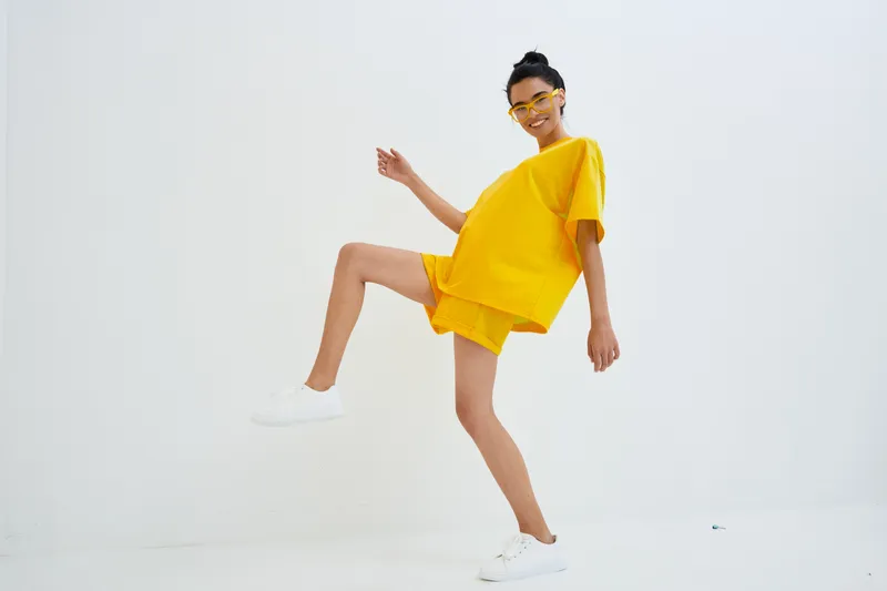 young woman in yellow outfit dancing energetically with white sneakers and sunglasses on white background, studio shot with bright casual style