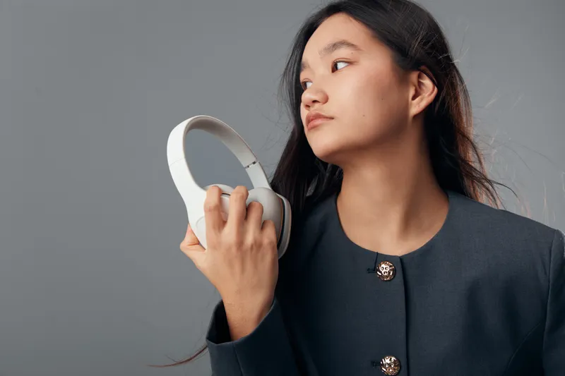 Young woman holding white headphones looking away on gray background. Fashionable female model with long black hair wearing dark jacket in studio for technology or music concept.