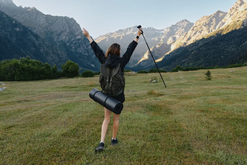 A hiker stands in a wide meadow with arms raised, wearing a backpack and gear. The bright sky and rugged mountains create a hopeful, adventurous outdoor scene.