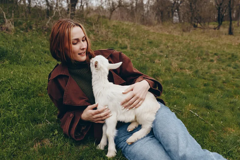 Relaxed woman lying on grass with a friendly goat kid in her lap, sharing a peaceful outdoor moment. Natural daylight, warm mood, close bond, lifestyle harmony, simplicity, carefree vibes, gentle