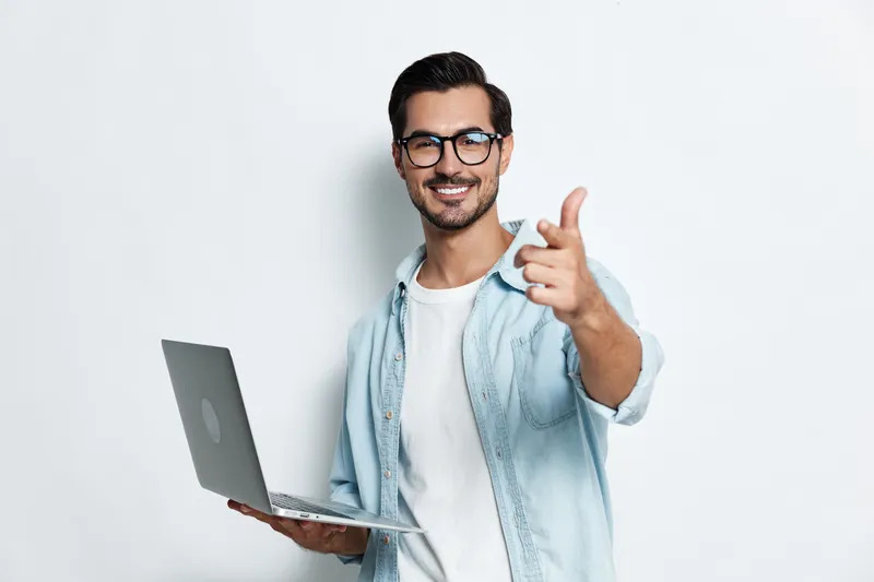 Smiling young man in glasses holding laptop and pointing finger at camera, dressed in casual outfit, standing against light background. Tech lifestyle concept.