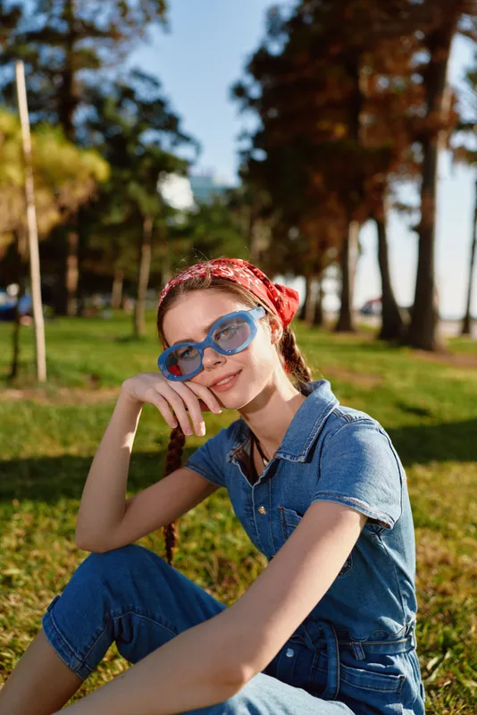 Lifestyle woman in modern boho-western denim street style wearing blue sunglasses and red bandana sitting outdoors with warm film color tones in a sunny park setting.