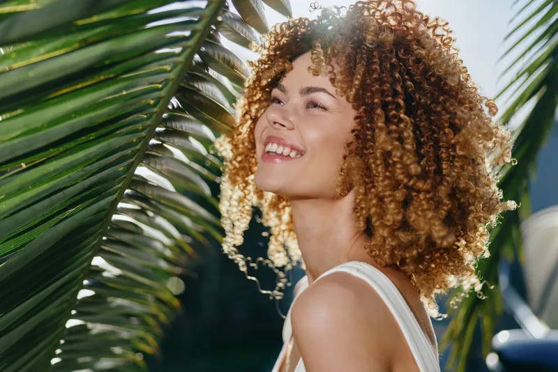 Smiling woman with curly hair enjoys sunny outdoor moment near tropical palm leaves, expressing happiness, natural beauty, and summer warmth in bright daylight.