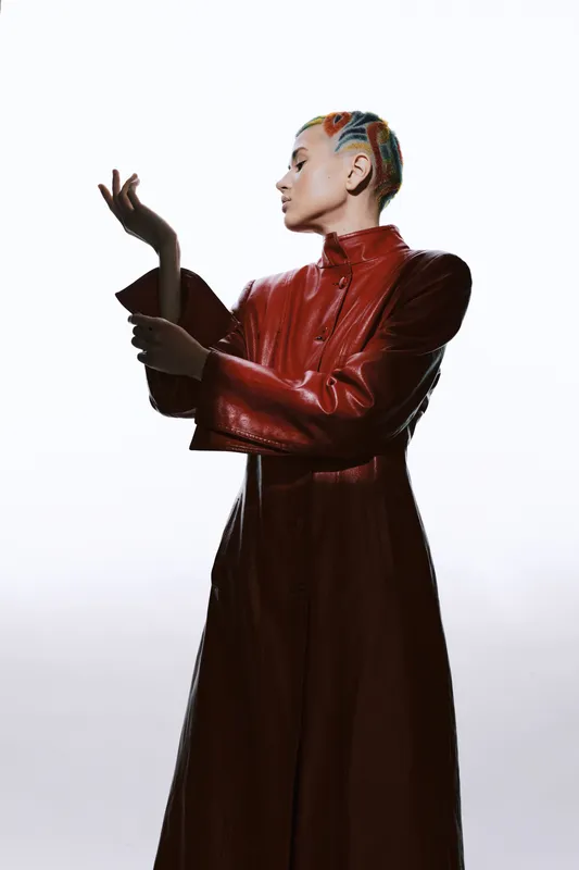 Woman with colorful braided hair wearing a long red leather coat poses stylishly against white background, highlighting fashion and modern aesthetics with artistic lighting.